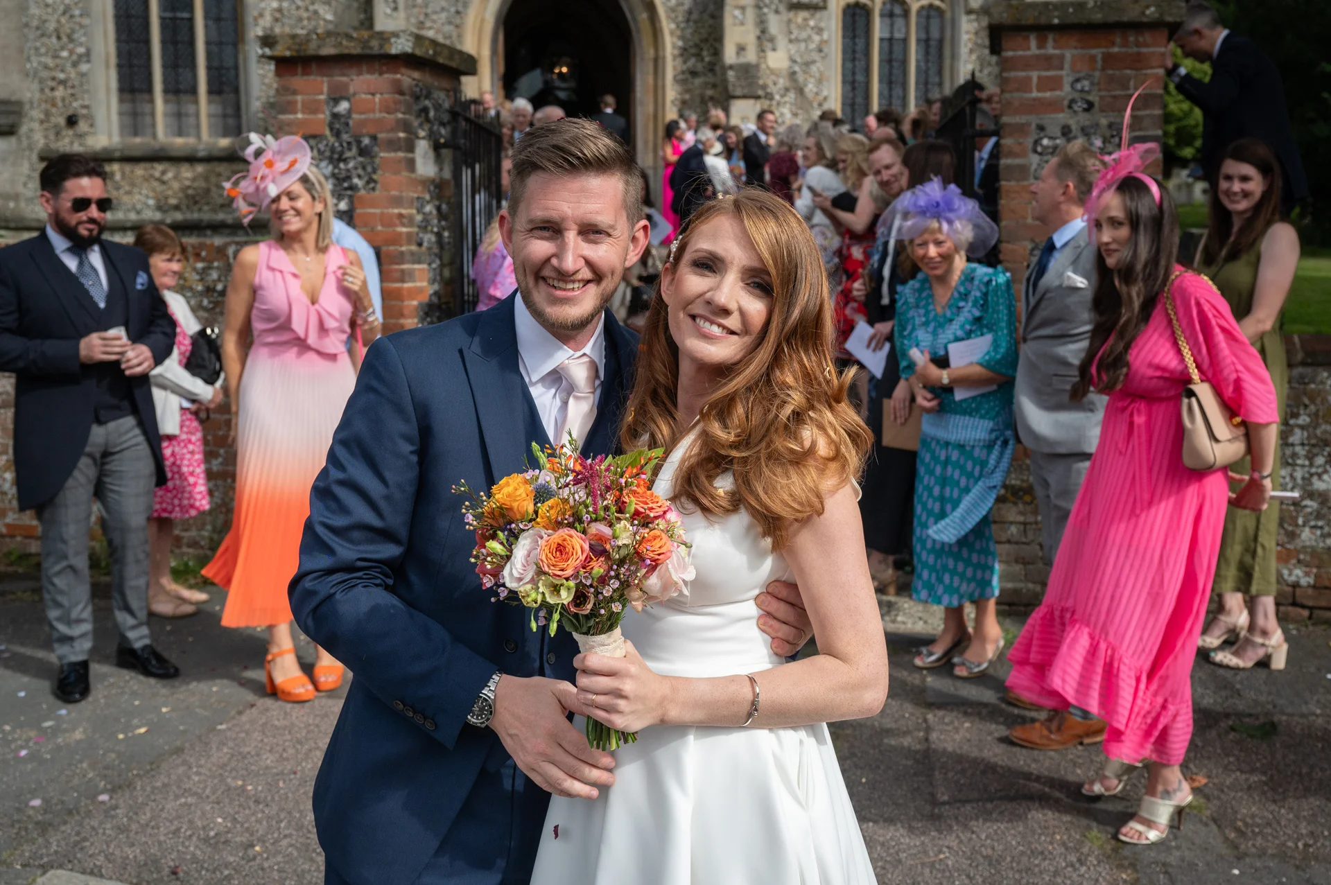 A smiling newlywed couple stand outside a traditional stone church holding a colourful bouquet, surrounded by wedding guests in the sunshine.