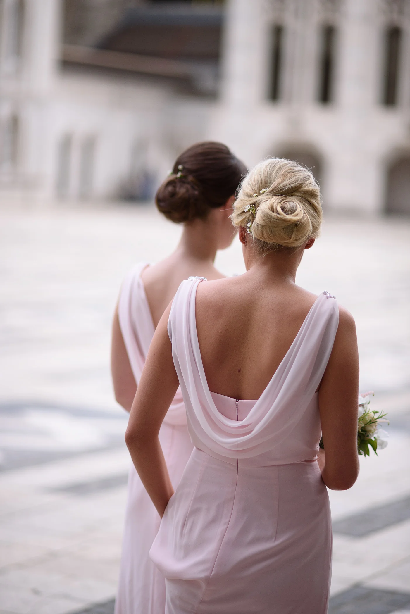 Two bridesmaids seen from behind wearing elegant pink dresses with draped cowls on the back and low buns adorned with small flowers.