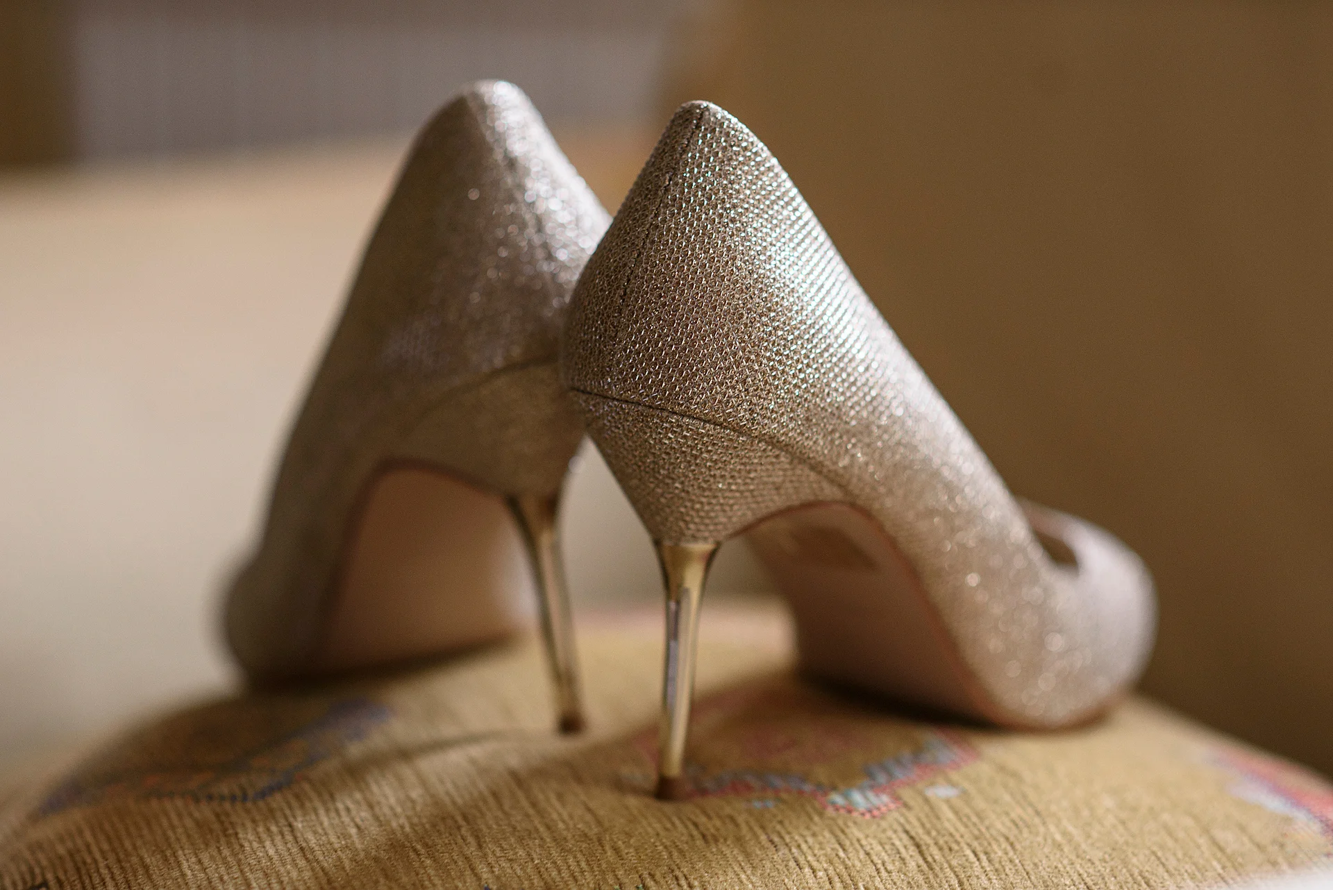 A close-up shot of sparkling gold stiletto heels resting on an upholstered surface during morning bridal preparations.