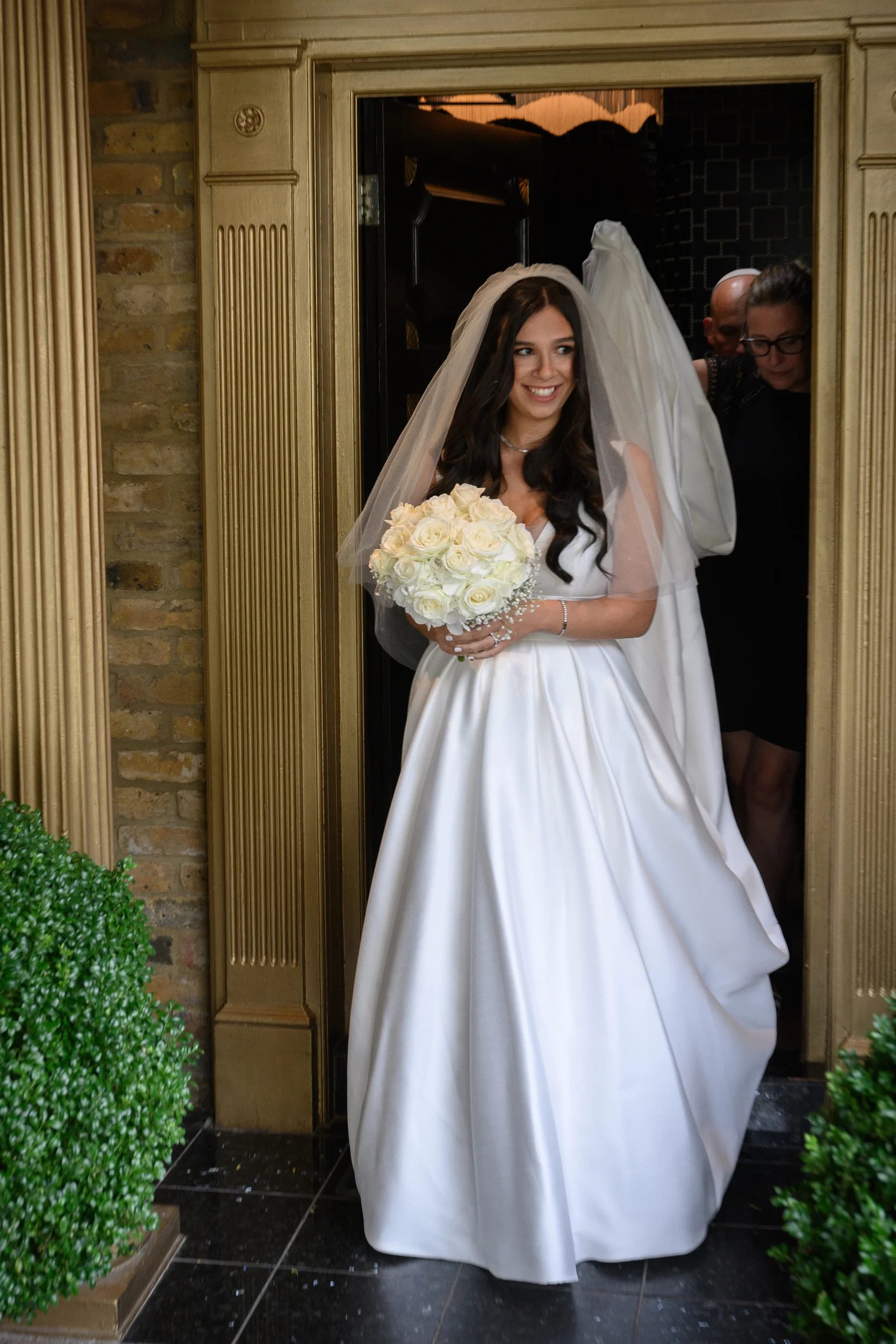 A smiling bride wearing a satin a-line gown and holding a bouquet of white roses steps through a golden doorway.
