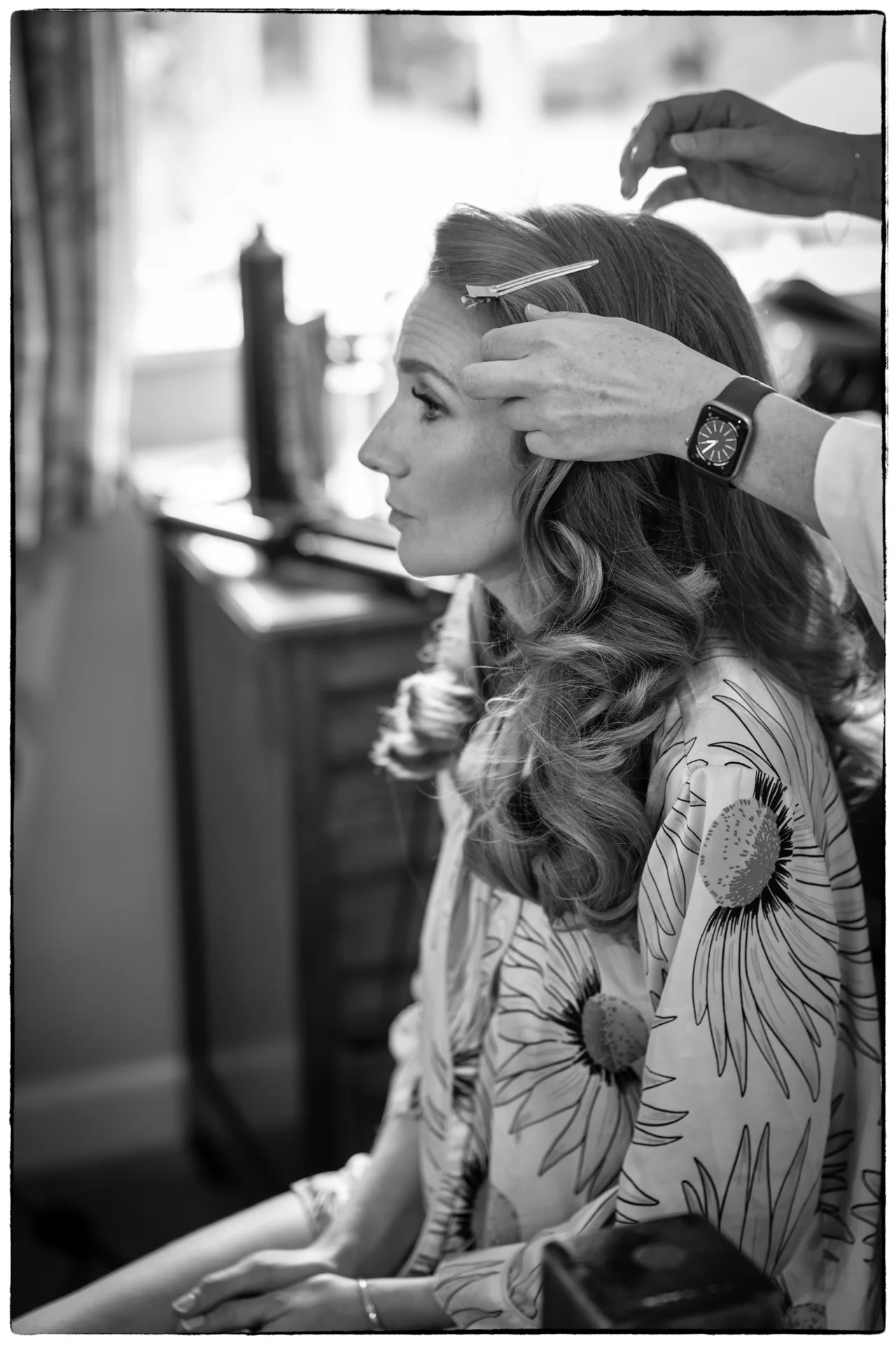 A black and white profile shot of a bride getting her hair styled into soft waves while wearing a floral robe during morning preparations.