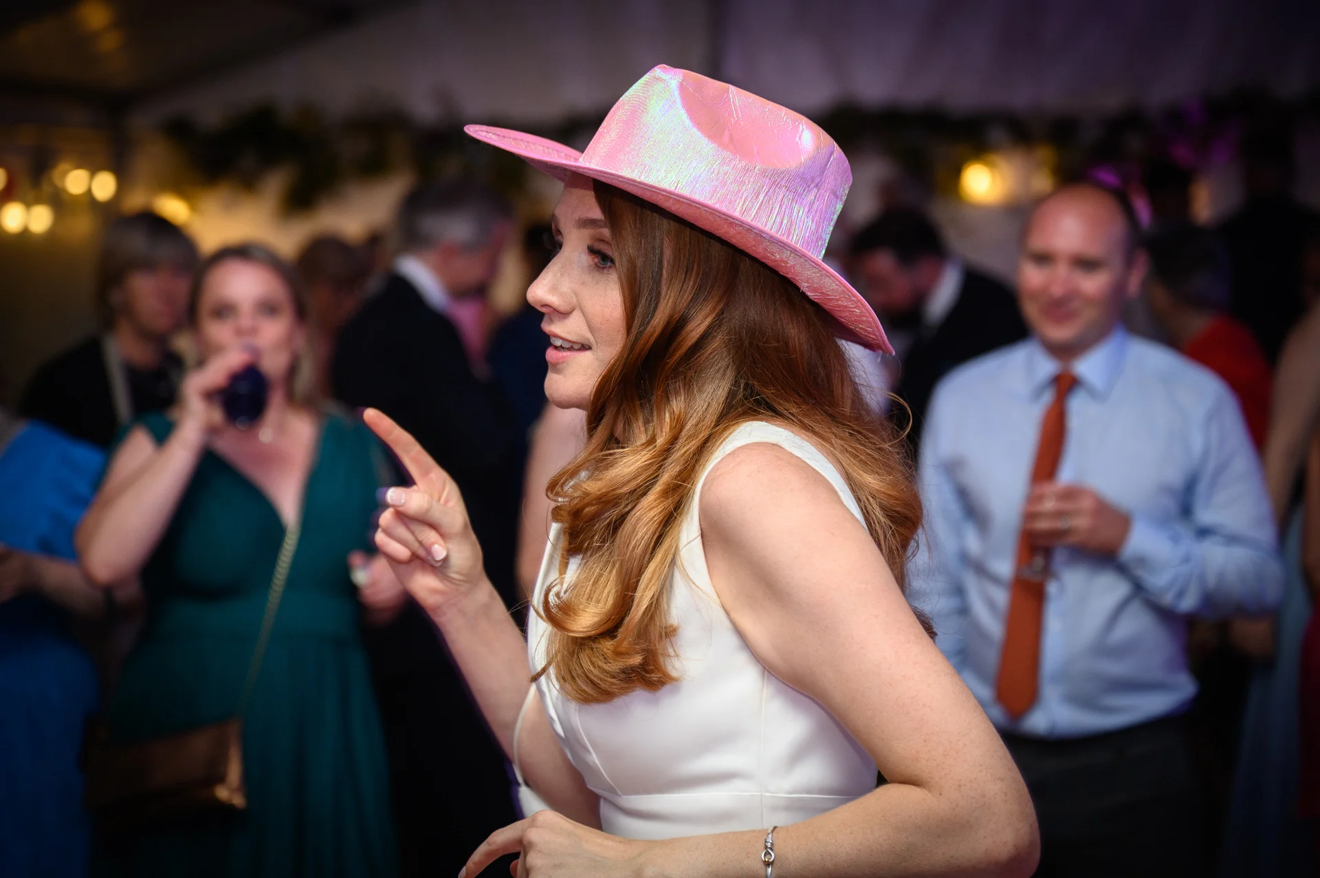 A guest at a lively evening wedding reception dances joyfully while wearing a shiny pink fedora hat and a white dress.