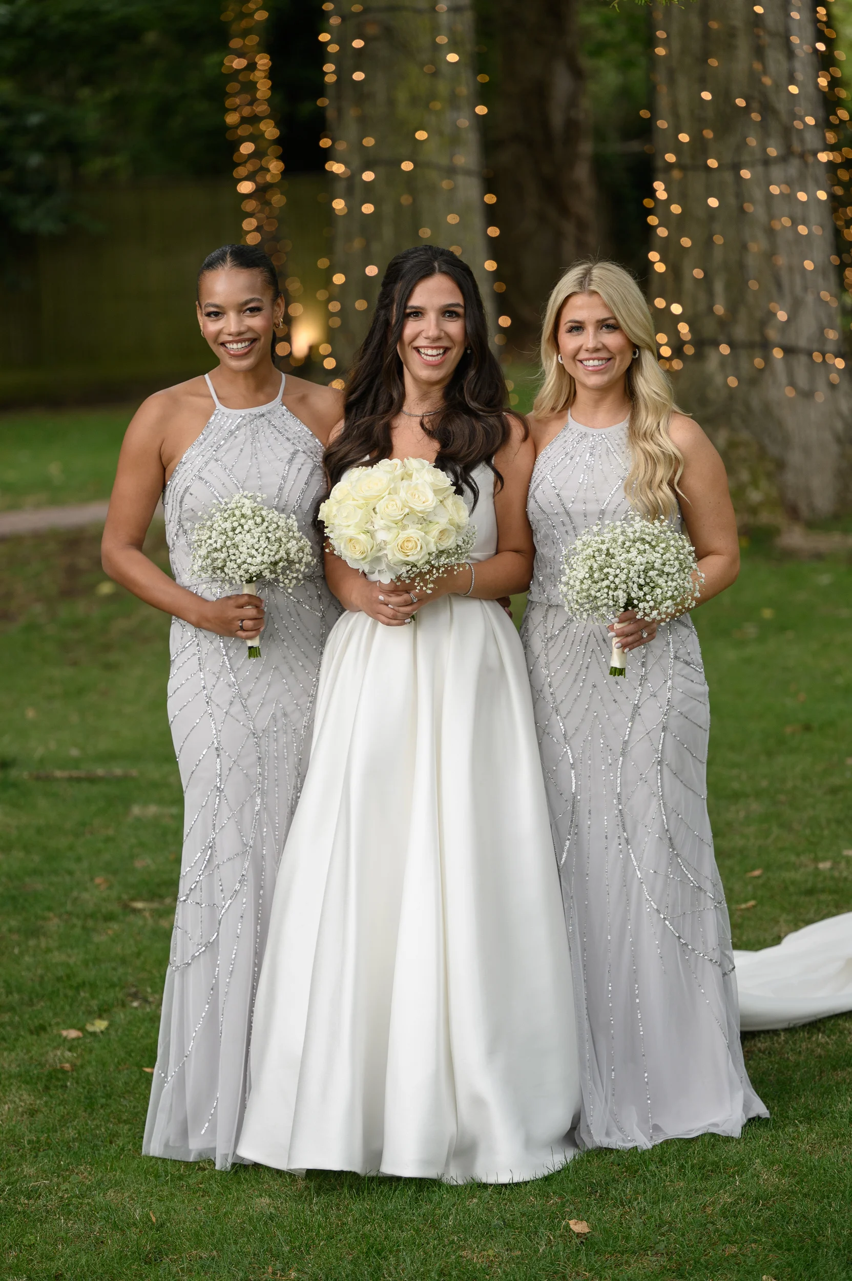 A smiling bride holding a white rose bouquet stands proudly outdoors flanked by two bridesmaids in sparkling grey gowns against a backdrop of twinkling fairy lights.