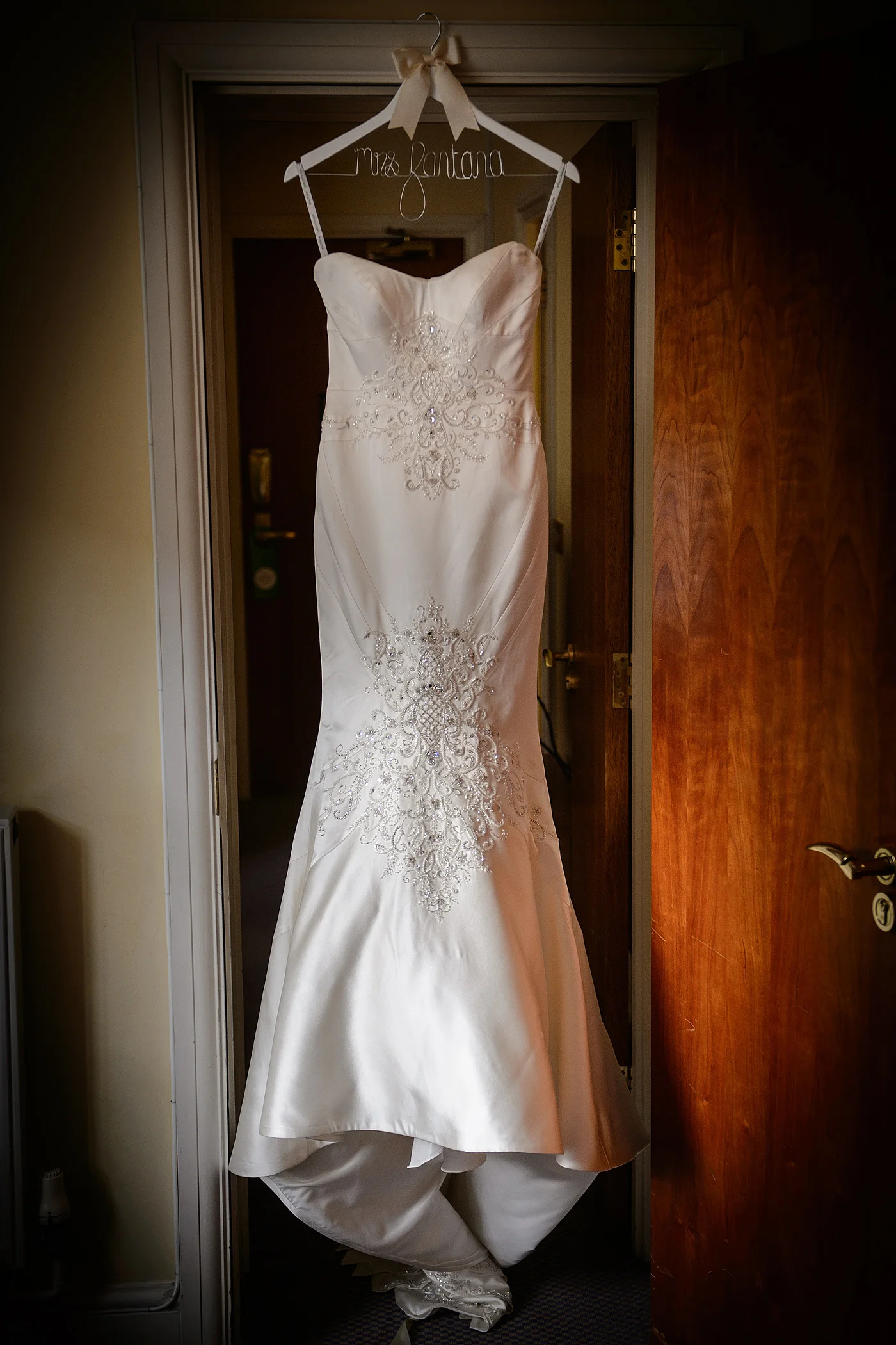 An intricately beaded and crystal-embellished strapless wedding dress hangs from a custom white hanger in an open wooden doorway.