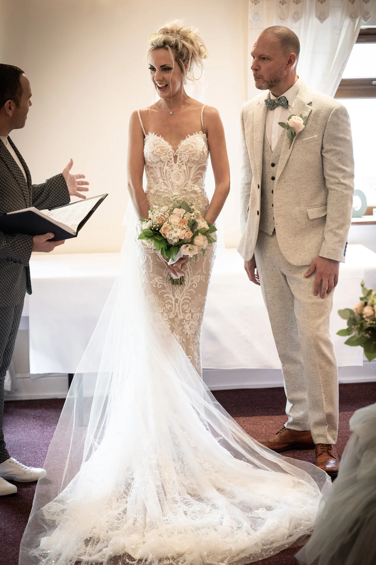 A laughing bride in a lace sheath dress with a long train and floral crown stands next to the groom in a light grey suit during their indoor wedding ceremony.
