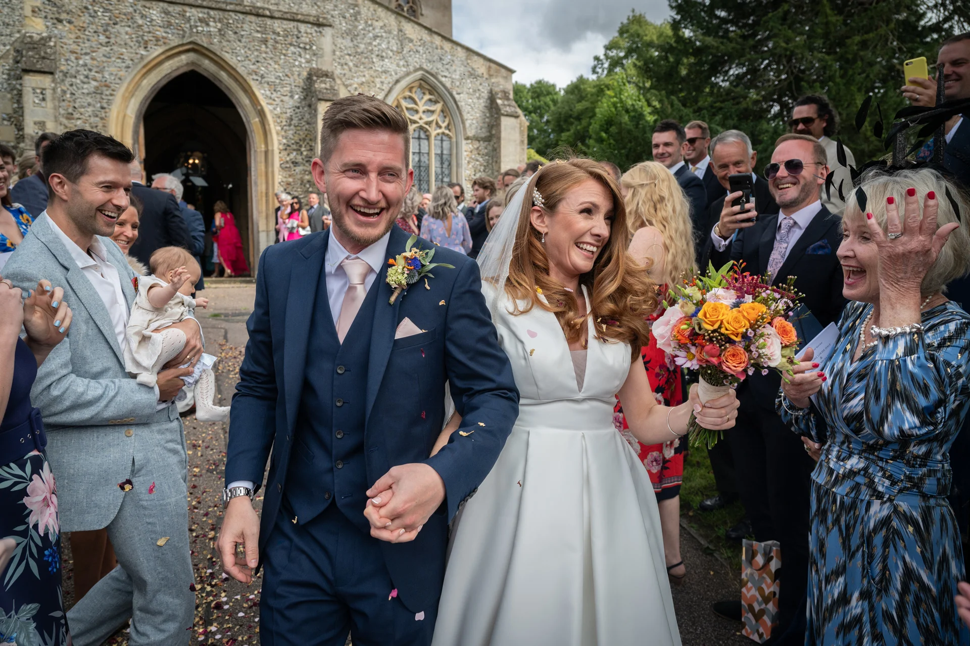A joyful newlywed couple walks hand in hand out of a stone church as guests cheer and toss confetti during their wedding ceremony exit.
