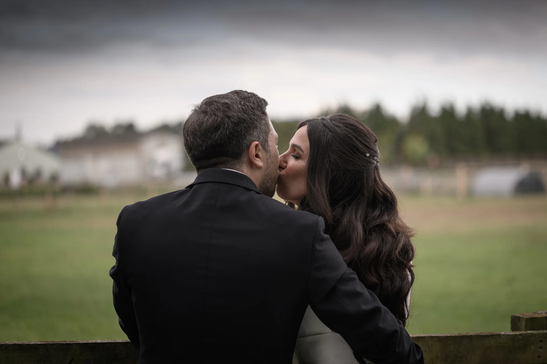 A newlywed couple share a tender kiss outdoors against a rural backdrop under an overcast sky.