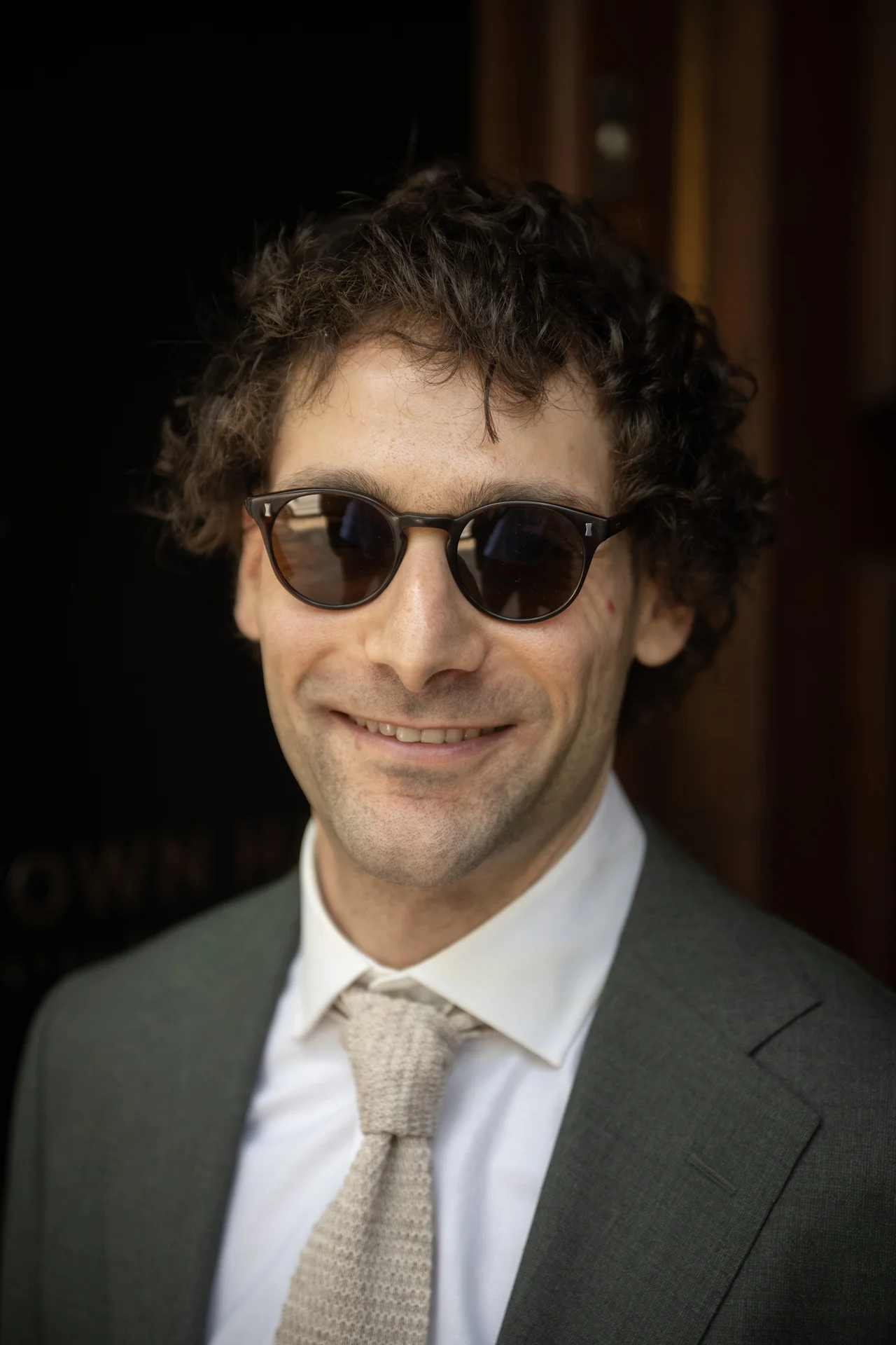 Close up portrait of a smiling groom with curly hair wearing dark round sunglasses, a grey suit, white shirt, and textured tie.
