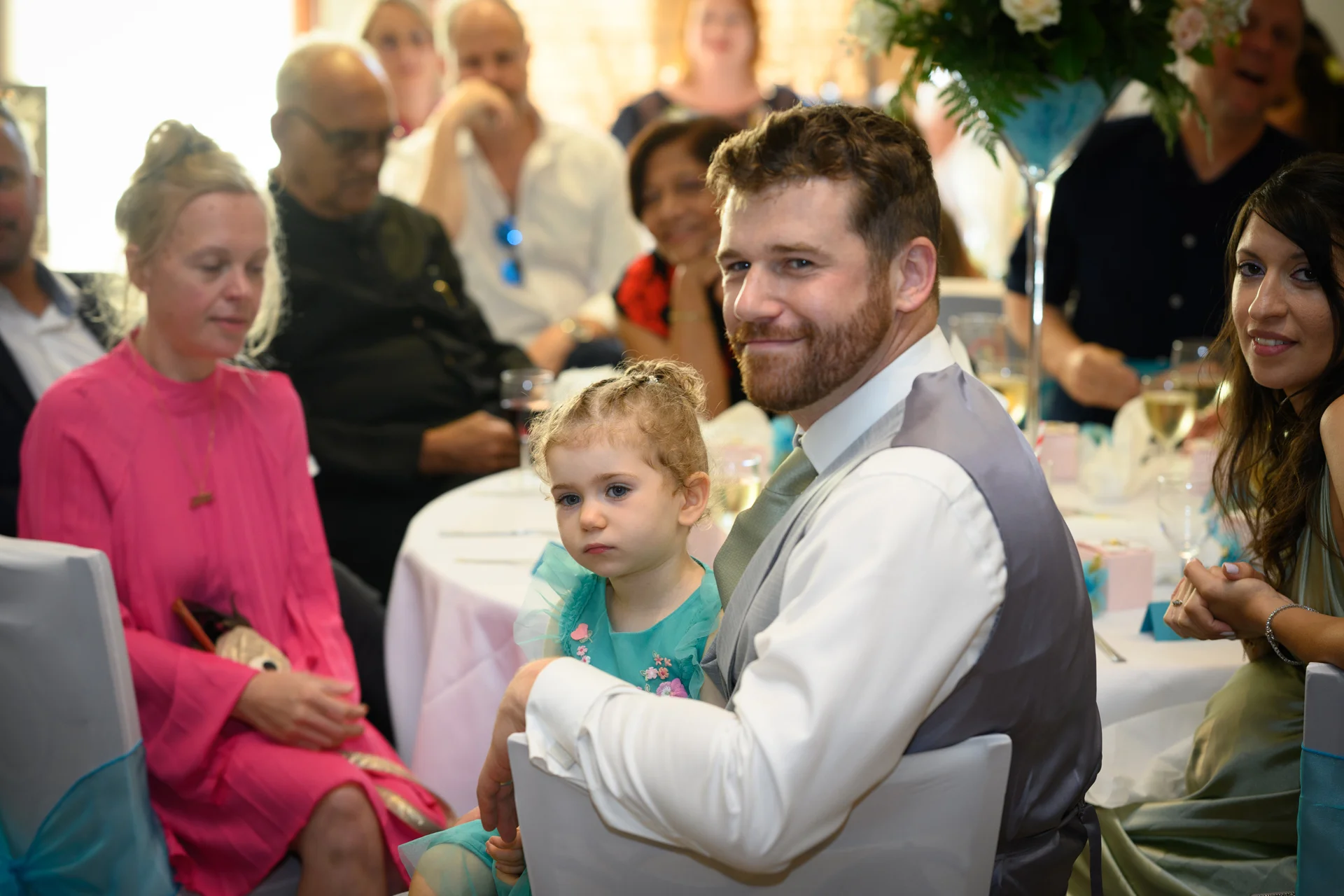 A bearded man in a white shirt and grey waistcoat sits at a wedding reception table holding a young girl on his lap, while guests look on.