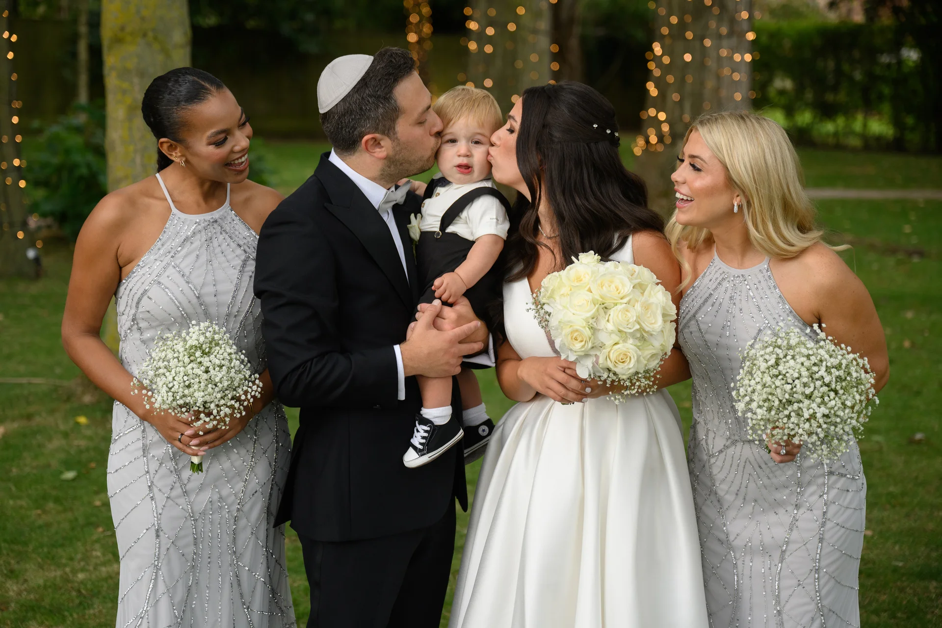 A groom wearing a kippah and a bride hold a young page boy while kissing his cheeks, flanked by smiling bridesmaids in embellished grey dresses holding bouquets of baby's breath.