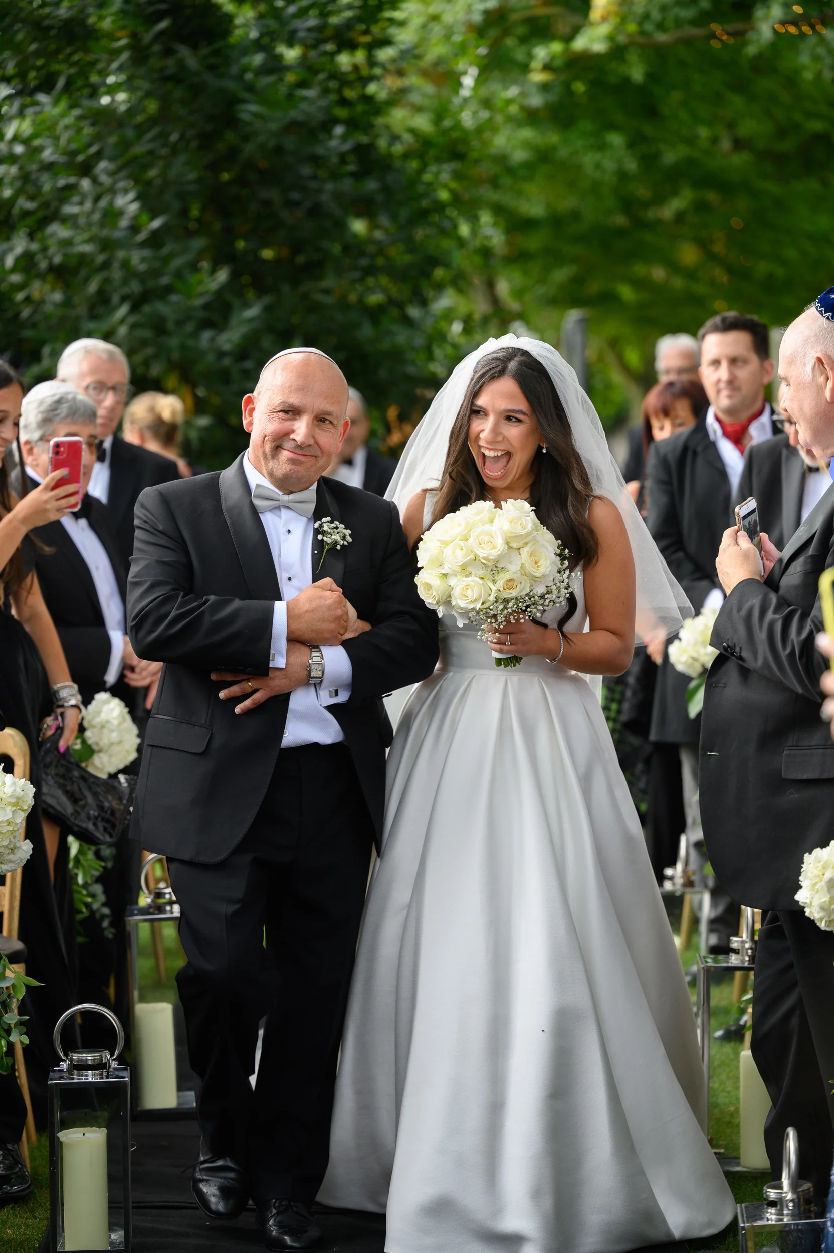A joyful bride laughing excitedly while walking down the outdoor aisle linked arm-in-arm with her father, holding a classic white rose bouquet.