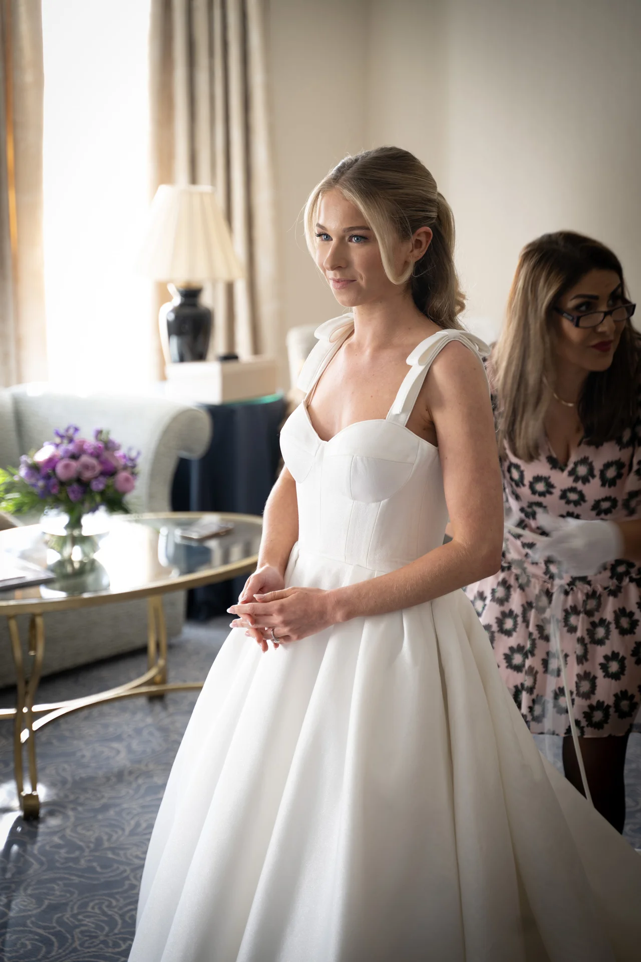 A beautiful bride stands smiling softly in her elegant white A-line wedding dress with sweetheart neckline while getting ready in a bright hotel room.