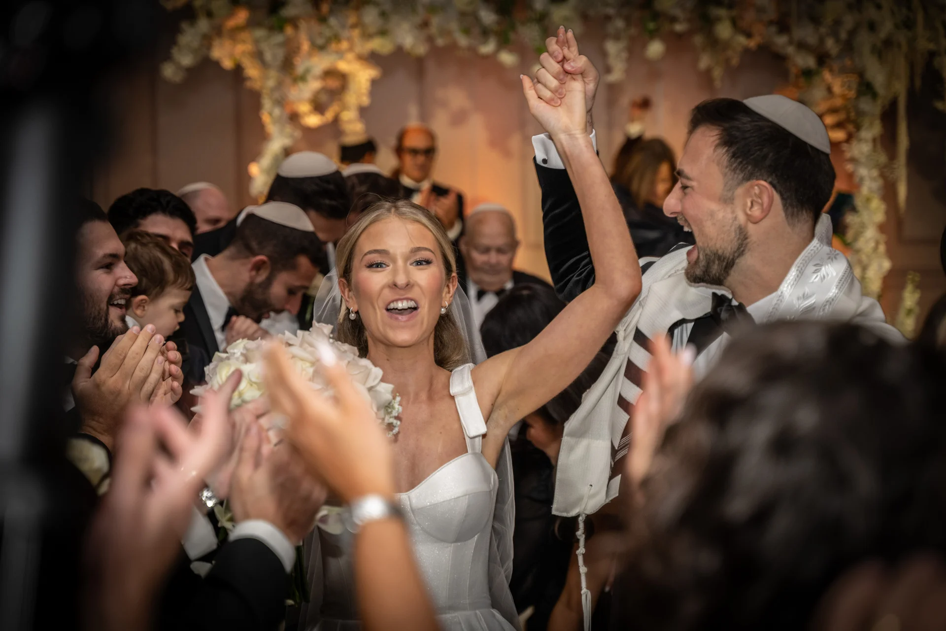 A joyous newlywed couple raises their hands in celebration as they walk back down the aisle surrounded by cheering guests at a Jewish wedding ceremony.