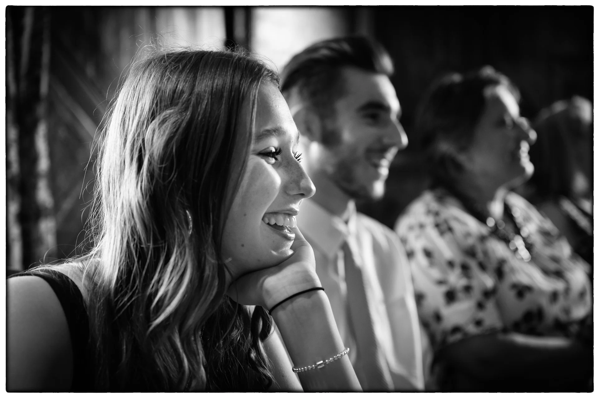 A smiling young female guest rests her chin on her hand in candid black and white while listening to wedding speeches.