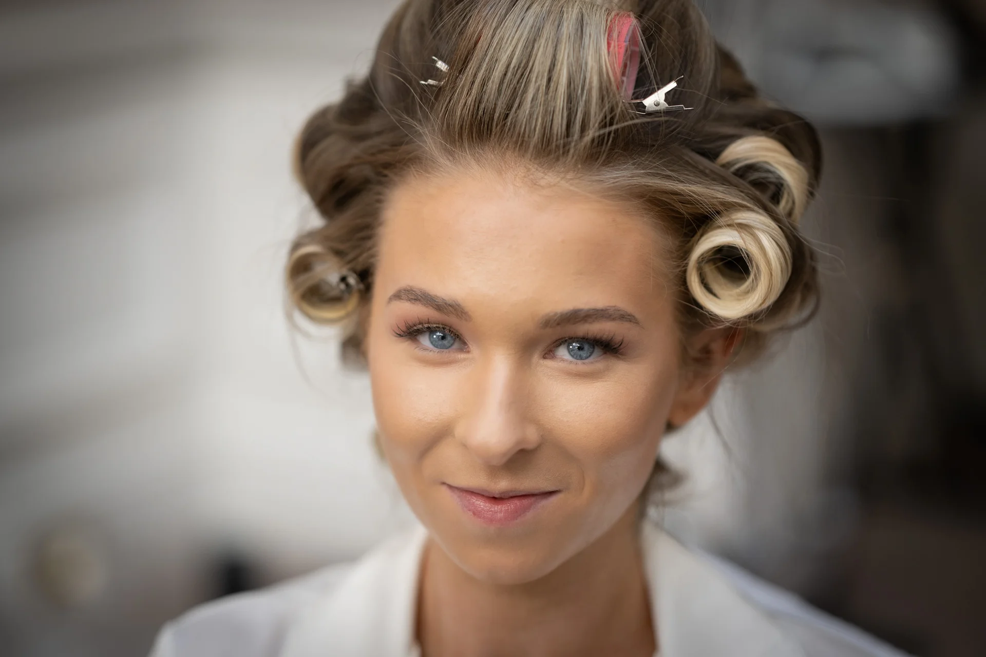 A close-up portrait of a bride with blue eyes smiling gently while wearing hair rollers and receiving morning beauty preparations.