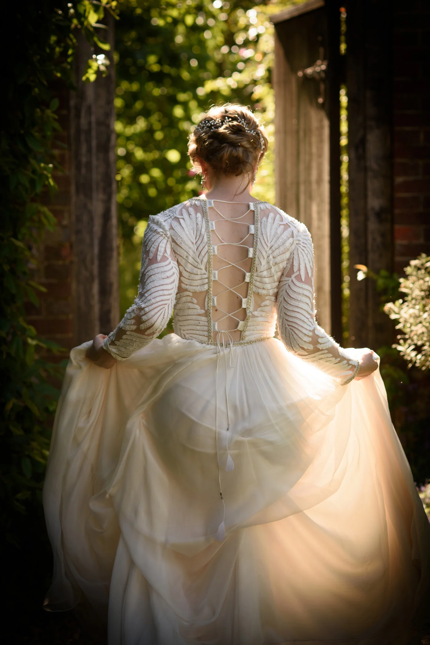 A bride viewed from behind twirls her ethereal gown in a sunlit garden doorway, highlighting the intricate lace-up back and sheer long sleeves.